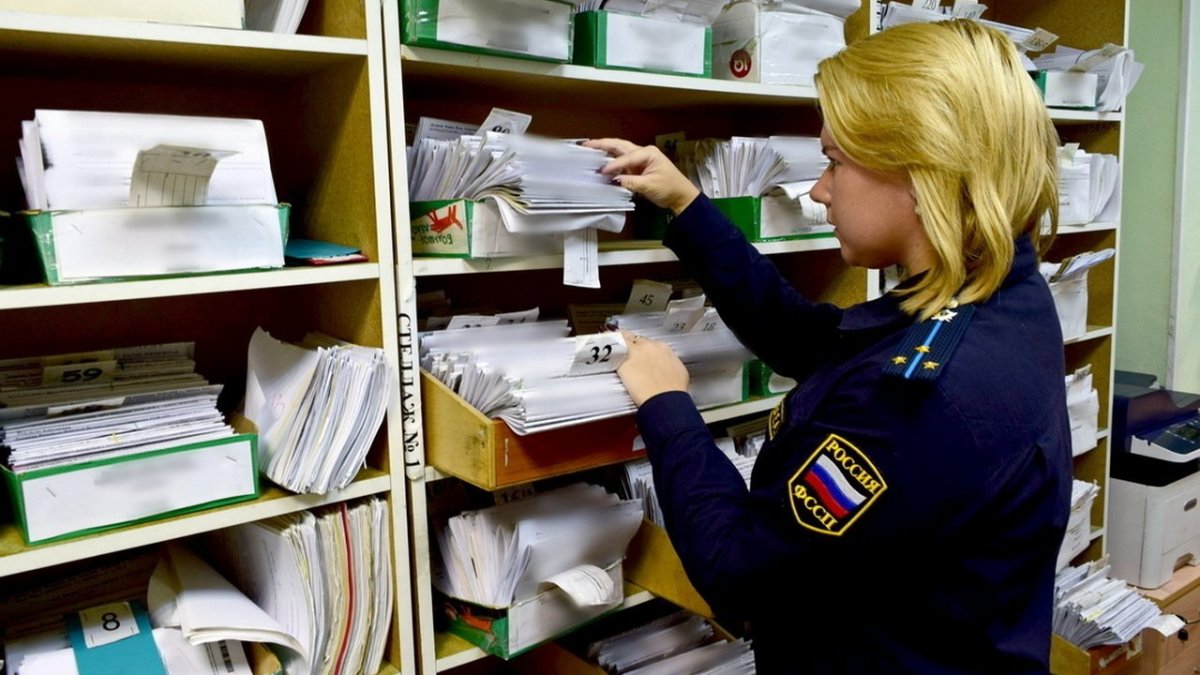 Женщина-судебный пристав стоит возле полок с документами.