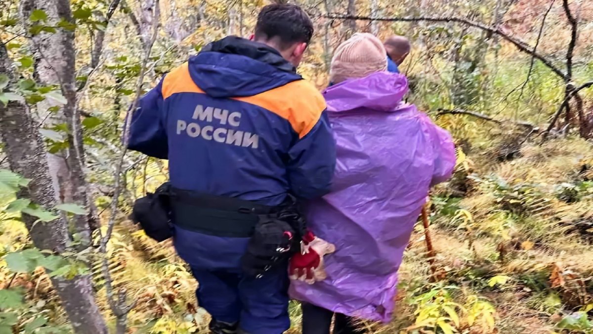 Спасатель ведет под руку пожилую женщину в лесу.