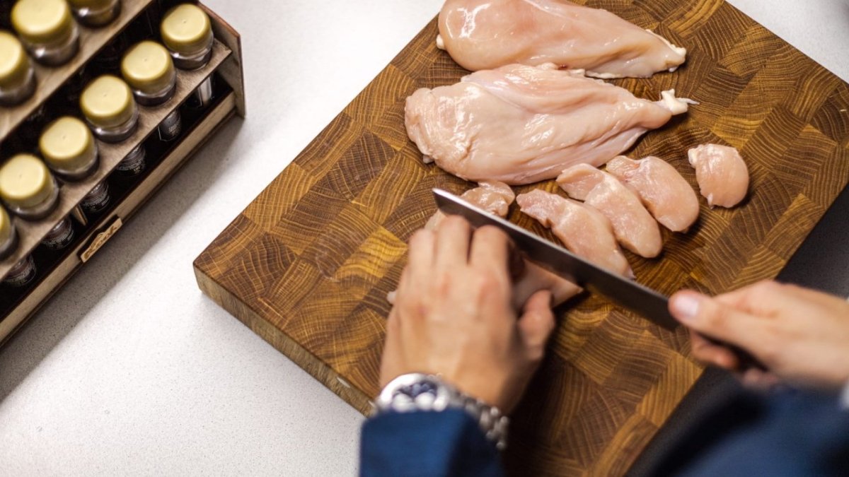 Мужчина ножом нарезает свежую куриную грудку на разделочной доске.